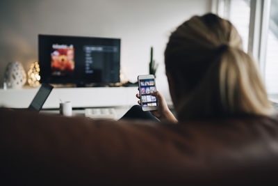 Rear view of teenage girl using smart phone app while watching tv in living room at home