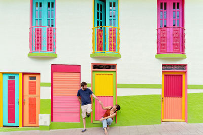Full body of happy young traveling couple holding hands and looking at each other near typical residential building with bright colorful doors and windows during holidays in colombia