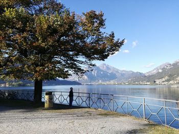 Man standing by railing by lake against blue sky