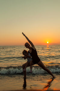 Mother with daughter doing yoga in sea during sunset