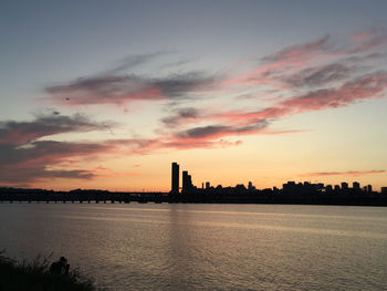 Silhouette buildings by river against sky during sunset