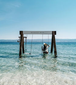 Rear view of young woman in summer clothes on sea swing on beach during summer vacation.