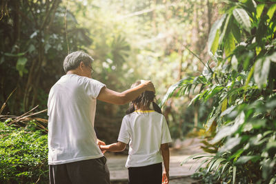 Rear view of grandfather with granddaughter walking amidst plants in forest