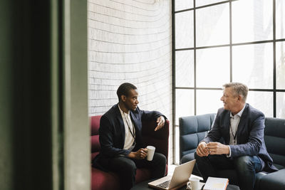 Male entrepreneur discussing with mature businessman sitting on sofa at convention center