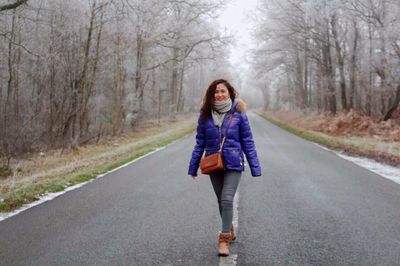 Portrait of woman in warm clothing walking on road amidst bare trees during winter