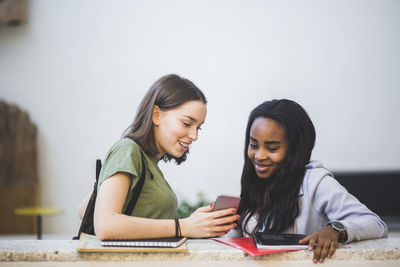 Smiling multi-ethnic female students sharing smart phone while standing in corridor at high school