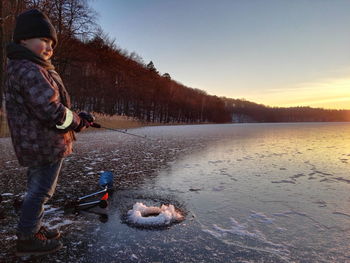 Side view of cute boy fishing in frozen lake during sunset