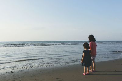 Rear view of sisters standing on shore at beach against clear sky