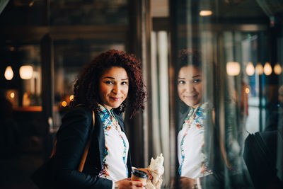 Portrait of smiling mid adult businesswoman holding food and drink standing by window in cafe