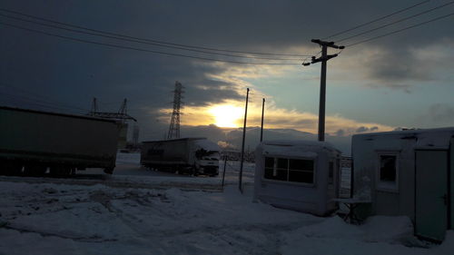 Snow covered electricity pylon against sky during sunset