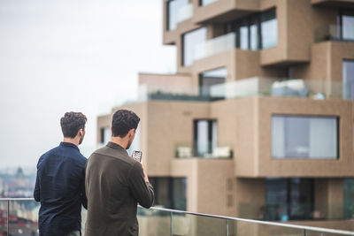 Man photographing through smart phone while standing by male friend on terrace