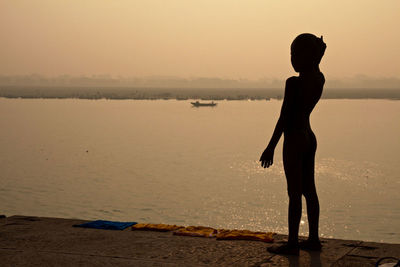 Silhouette boy standing at ganges riverbank against sky