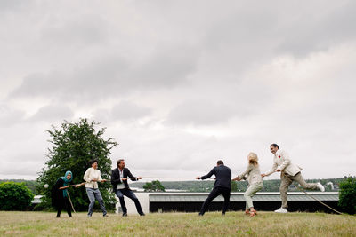 Full length of businessmen and businesswomen playing tug-of-war in lawn against sky