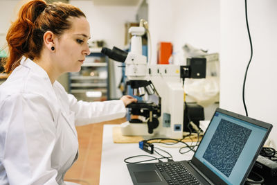 Side view of concentrated female chemist looking at screen of netbook with enlarged image while examining chemical sample in microscope during work in laboratory