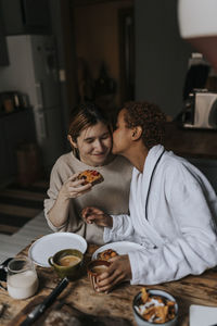 Non-binary person kissing friend while having breakfast at home