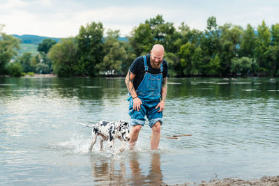 Happy man with beard in casual clothes playing with puppy while standing in lake water in summer