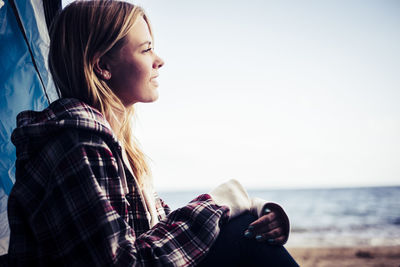 Side view of young woman looking away while sitting at beach against sky