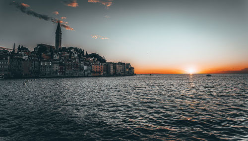 Sea by buildings against sky during sunset