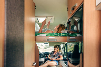Woman lying on camper van bunk bed reading book while her friend looking mobile