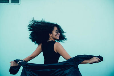 Back view of happy young ethnic woman with afro curls wearing black dress standing and twisting in city street near building wall in daylight