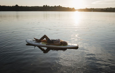 High angle view of woman lying on paddleboard in lake against sky during sunset