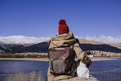 Rear view of woman sitting with dog on rock by lake during winter