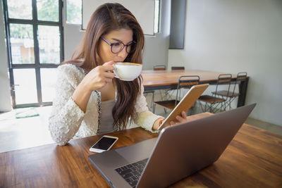 Businesswoman drinking coffee while using technologies at desk in cafe