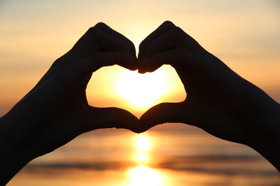 Close-up of hand making heart shape against sky during sunset