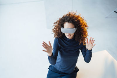 Young businesswoman with curly hair wearing virtual reality simulator sitting on desk in office