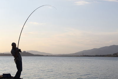 Man fishing in lake against sky during sunset