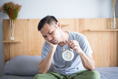 Sleepy man holding alarm clock while sitting on bed at home
