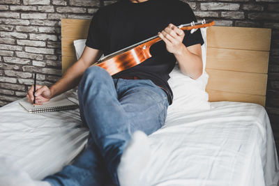 Low section of musician writing on sheet music while sitting with ukulele on bed at home