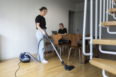 Smiling woman cleaning hardwood floor with vacuum cleaner near girlfriend using laptop at home