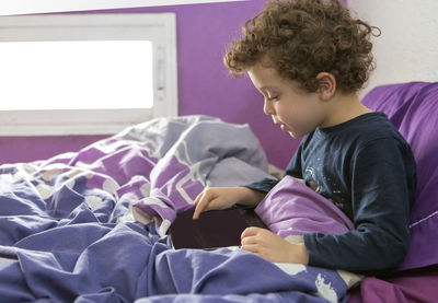 Child entertaining himself in bed in the morning with a tablet resting on the pillow 