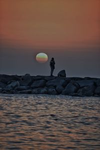 Man standing on rock by sea against sky during sunset