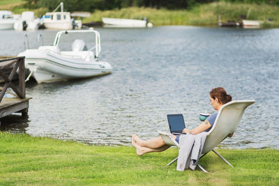 Full length side view of mature woman using laptop on deck chair at lakeshore