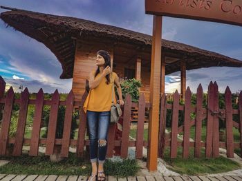 Full length of young woman looking away while standing by fence against cloudy sky during sunset