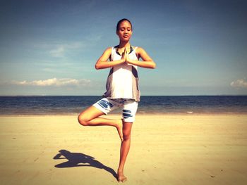 Full length of woman doing yoga in tree position at beach against sky