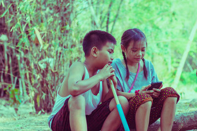 Girl using phone by brother sitting on fallen tree