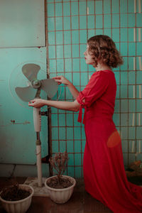 Side view of young woman adjusting electric fan against wall