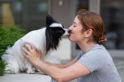 Caucasian red-haired woman kissing pappilion dog outdoors. black and white continental spaniel