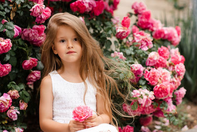 Cute baby girl 5-6 year old hold rose flower sit over bloom bushes in garden outdoor. spring season