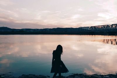 Silhouette girl standing by lake against sky during sunset