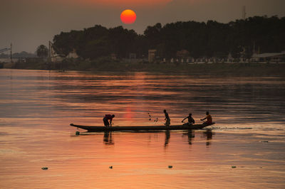 People fishing on river against trees during sunset