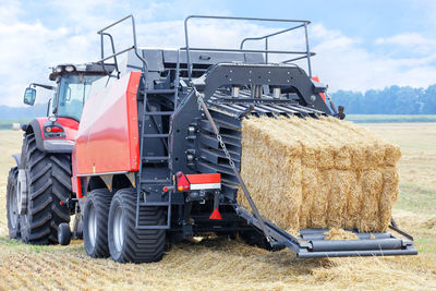 An agricultural tractor with a trailer forms bales of straw into dense briquettes in a wheat field.