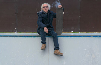 Full length portrait of mature man holding skateboard while smoking cigarette