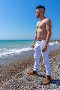 Full length of shirtless man standing at beach against clear sky