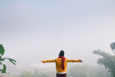 Rear view of woman with arms outstretched standing against landscape and sky during foggy weather