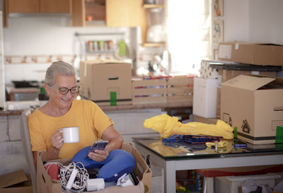 Senior woman drinking coffee cup while using mobile at home