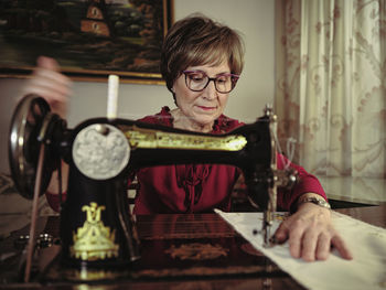 Senior lady in glasses using retro sewing machine to create linen napkin in cozy room at home